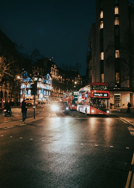 A nighttime view of a city street with illuminated buildings and trees along the sidewalk. In the foreground, the road surface appears wet, reflecting the glow of streetlights. A red double-decker bus